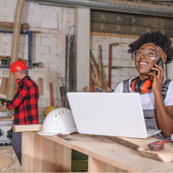 Business owner speaks on the phone while working on a laptop computer in her small wood shop.